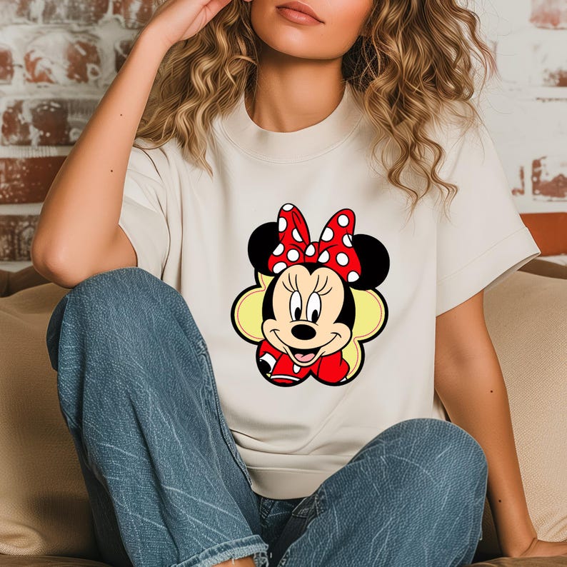 May include: A cream-colored t-shirt featuring a colorful graphic of Minnie Mouse. Minnie Mouse is depicted with a red bow with white polka dots, black ears, and a red dress. The graphic is centered on the shirt.