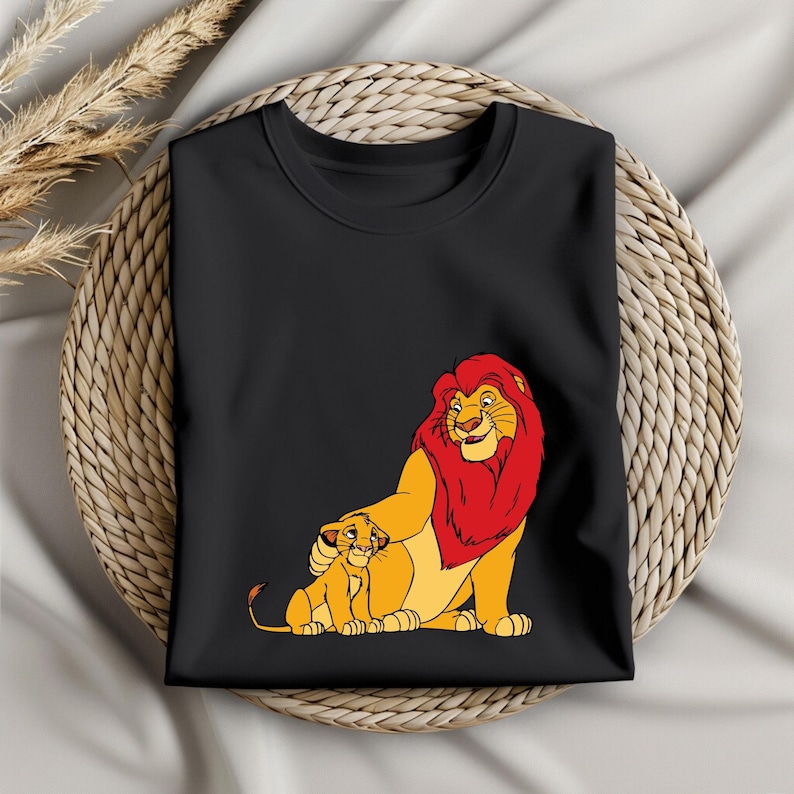 May include: Black t-shirt featuring a cartoon illustration of a lion and cub. The lion has a red mane and the cub is yellow. The design is printed on a folded black t-shirt, likely made of cotton or a similar fabric.