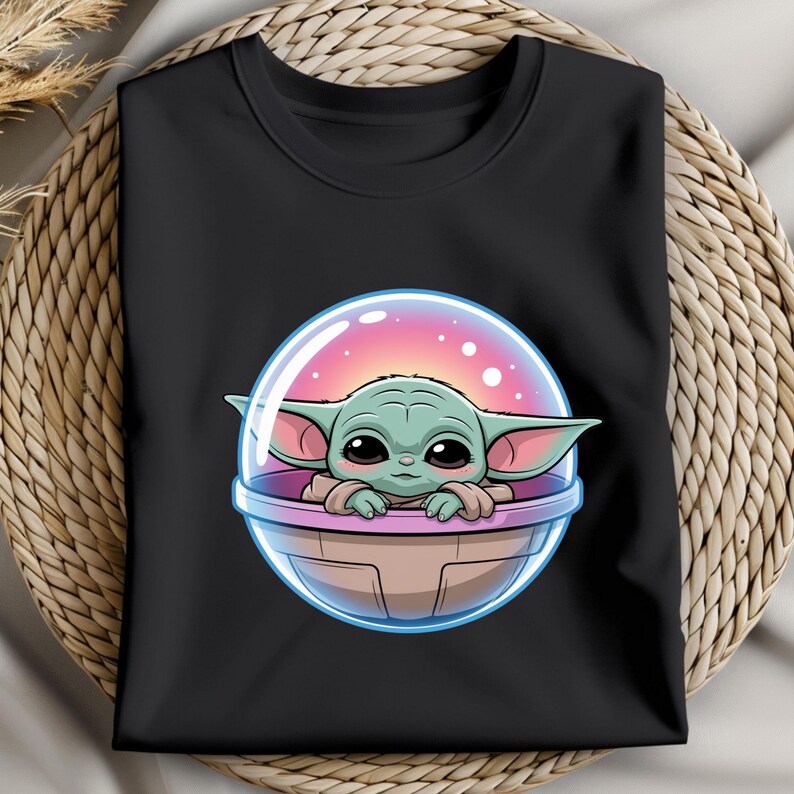 May include: Black long-sleeve shirt featuring a cartoon illustration of a green alien-like character in a clear bubble. The character has large eyes and ears, with a pink and purple background inside the bubble.