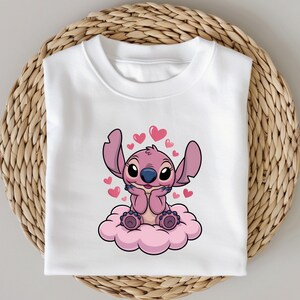 May include: White sweatshirt featuring a cartoon character with pink fur, large ears, and a blue nose, sitting on a pink cloud. The design includes pink hearts around the character. The sweatshirt is folded on a woven surface.