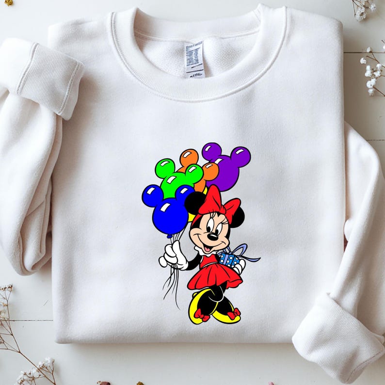 May include: White sweatshirt featuring a colorful Minnie Mouse graphic. Minnie Mouse is holding a bunch of balloons and a gift. The balloons are blue, green, orange, and purple. Minnie is wearing a red dress and bow.
