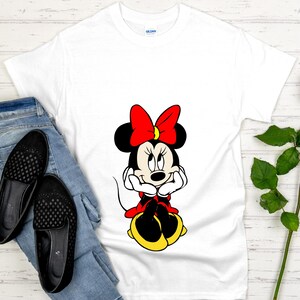 May include: White t-shirt featuring a cartoon image of Minnie Mouse. Minnie is depicted with a red bow, black ears, and a red and yellow outfit. The shirt is paired with blue jeans and black shoes.