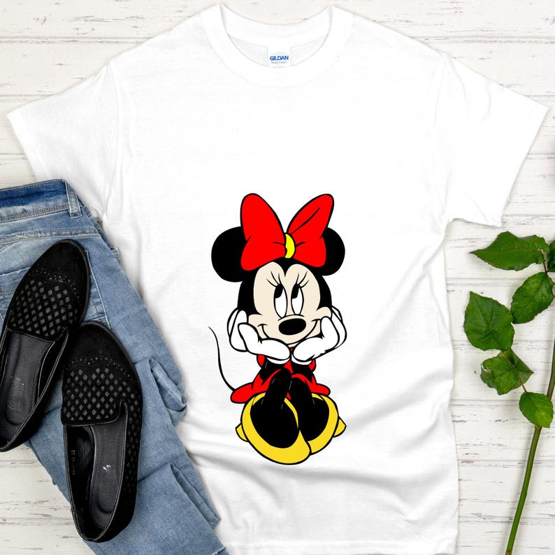 May include: White t-shirt featuring a cartoon image of Minnie Mouse. Minnie is depicted with a red bow, black ears, and a red and yellow outfit. The shirt is paired with blue jeans and black shoes.