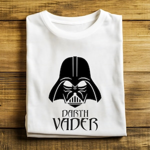 May include: White t-shirt featuring a black Darth Vader graphic. The design includes Vader's helmet and the text "Darth Vader" in a stylized font. The shirt is folded neatly on a wooden surface.