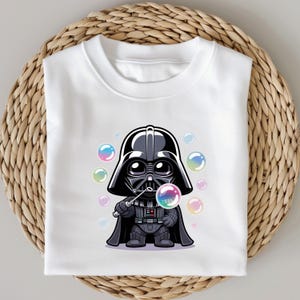 May include: White t-shirt featuring a cartoon Darth Vader character blowing bubbles. The character is depicted in black and gray with a bubble wand and colorful bubbles. The shirt is folded on a woven surface.