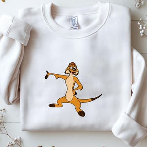 May include: White sweatshirt featuring a cartoon meerkat character in shades of brown and tan. The meerkat is in a dynamic pose with one arm extended and the other on its hip. The sweatshirt has long sleeves and a crew neck.