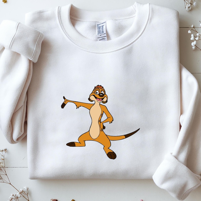 May include: White sweatshirt featuring a cartoon meerkat character in shades of brown and tan. The meerkat is in a dynamic pose with one arm extended and the other on its hip. The sweatshirt has long sleeves and a crew neck.
