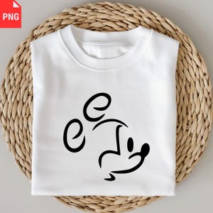 May include: White sweatshirt featuring a black minimalist Mickey Mouse design. The graphic is a simple outline of the character's face and ears. The sweatshirt is folded and placed on a woven, light brown surface.