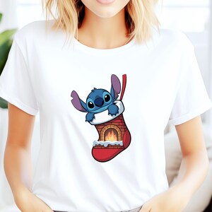 Pu&ograve; includere: T-shirt bianca con un'immagine a cartoni animati di Stitch di Lilo & Stitch. Stitch, un alieno blu, &egrave; all'interno di una calza di Natale rossa con un design a camino. La calza ha un polsino bianco e un nastro rosso.