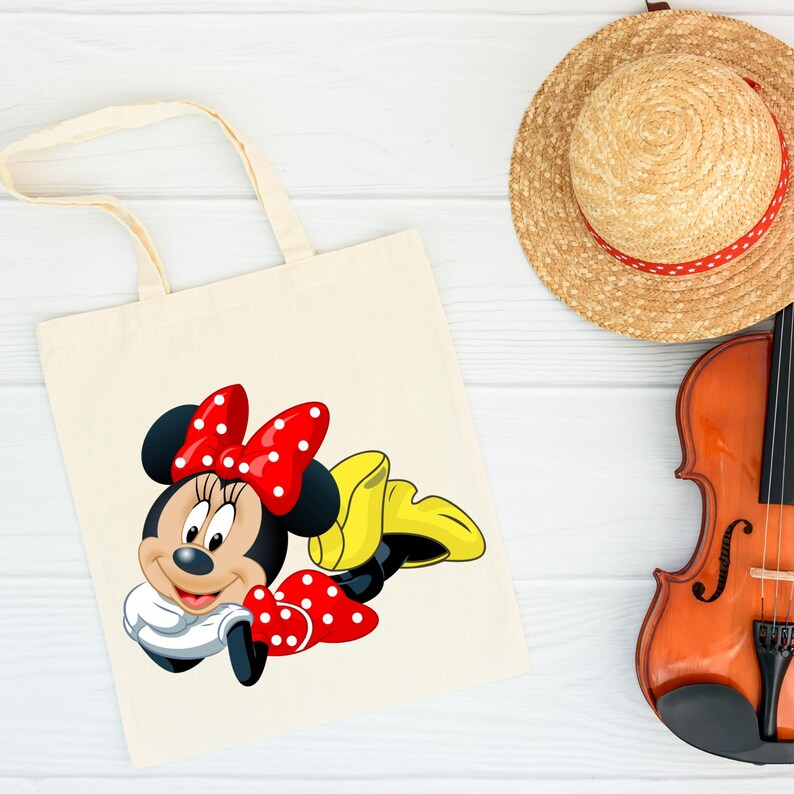 May include: A cream-colored tote bag featuring a cartoon illustration of Minnie Mouse. Minnie Mouse is depicted with a red bow with white polka dots, a white shirt, and yellow shoes. A straw hat with a red ribbon and a violin are also in the image.
