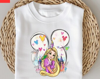 May include: White sweatshirt featuring a colorful illustration of a princess with long blonde hair, a green chameleon, and a castle. The design is set within a Mickey Mouse head outline with balloon and firework accents. A whimsical design.