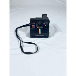 Peut inclure: Un appareil photo Polaroid Land noir avec une bande arc-en-ciel et les mots "Time-Zero OneStep" et "Polaroid Land Camera" imprimés sur le devant. L'appareil photo a une sangle noire attachée. L'appareil photo est sur fond blanc.