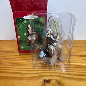 Puede incluir: Un adorno navideño de Star Wars con un personaje con un sable de luz verde, empaquetado en una caja roja Hallmark. El adorno está en una funda de plástico transparente. La caja tiene una imagen del personaje con un sable de luz.