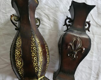 May include: Two decorative brown vases with ornate details. One vase features a gold-colored, patterned design, while the other has a fleur-de-lis emblem. Both vases have handles and a distressed finish.