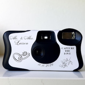 Pode incluir: Câmera descartável em preto e branco com o texto "Mr. & Mrs. Larson" e "Capture the Love". Apresenta um gráfico de anéis de casamento e a data 11.11.2026. Ideal para lembrancinhas de casamento ou para capturar memórias.