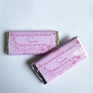 May include: Two pink chocolate bars with silver foil wrapping. The wrappers feature the text "MY DAY WOULD BE SO MUCH Sweeter WITH YOU BY MY SIDE" in a decorative font, along with small illustrations of swans, hearts, and other designs.