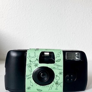 May include: Black disposable camera with a light green paper band featuring illustrations of cocktails, bikinis, and flowers. The band has the text "Flash Me" and "Bikinis Martinis" with the date 04.11.2026.