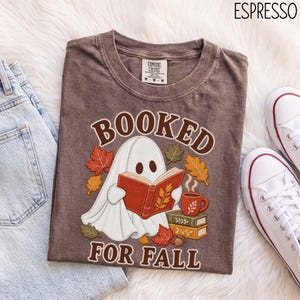 May include: An espresso-coloured t-shirt featuring a graphic of a white ghost reading a red book, surrounded by autumn leaves, a stack of books, and a mug. The text reads BOOKED FOR FALL. The shirt has a crew neck and a Comfort Colors brand label.