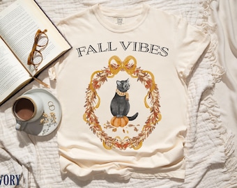 May include: Ivory-colored t-shirt with the words "FALL VIBES" above a watercolor illustration of a black cat wearing glasses and a scarf, sitting on a pumpkin within a fall wreath.