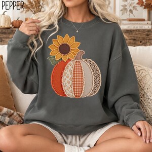 May include: A charcoal gray crewneck sweatshirt featuring a large graphic of a sunflower and a pumpkin. The pumpkin is composed of vertical fabric panels with various patterns including floral, gingham, and leopard print in orange and beige.