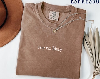 May include: Espresso-colored t-shirt with the phrase "me no likey" in white. The shirt has a crew neck and a small tag near the collar. A silver necklace with a pendant is draped across the shirt. A brown hat is visible.