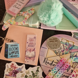 May include: A collection of stationery and book-related accessories. The image shows a bookmark, a keychain, a pom-pom, a notepad with the text "DO NOT DISTURB PLEASE", and decorative stickers. The colour scheme features pastels and bright shades.
