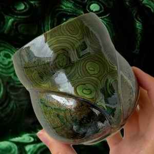 May include: A handcrafted, glossy ceramic bowl with a unique, organic shape. The bowl features a swirling, concentric pattern in shades of green and brown, reminiscent of natural stone. The interior reflects light, enhancing the depth of the design.