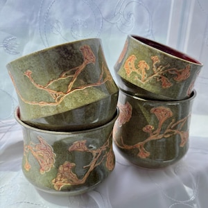 May include: A stack of four ceramic bowls. The bowls are a muted green with a reddish-orange floral design. The interior of one bowl is a deep red. The bowls are sitting on a white surface.