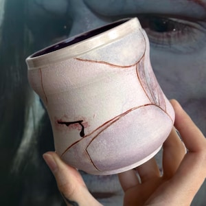 May include: A handmade ceramic cup with a unique design. The cup is primarily white with a purple interior and brown accent lines. A dark red, stylized blood drop is painted on the side. The cup is held in a hand.