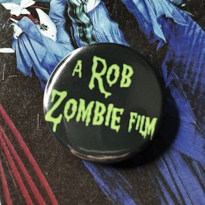 May include: A black button with the text "A ROB ZOMBIE FILM" in neon green. The button is set against a backdrop of a movie poster with figures in blue and black.