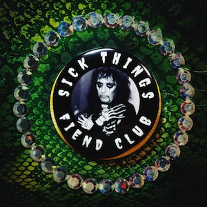 May include: A black button with the text "SICK THINGS FIEND CLUB" encircling a black and white image of a person with skeleton-themed makeup. The button is surrounded by a circle of iridescent beads, set against a green background.