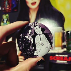 May include: A monochrome button badge showing a vintage image of a woman in a black dress and a man in a suit. The badge is held in front of a blurred background with a woman with dark hair and a red lip.