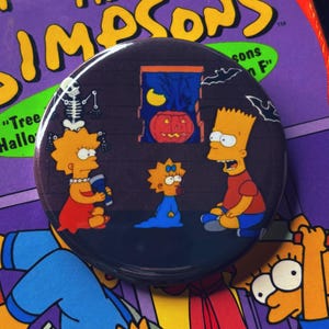 May include: A round button featuring The Simpsons characters Lisa, Maggie, and Bart, with a Halloween theme. The button has a dark background with a jack-o'-lantern, moon, and bats. Lisa is holding a blue object, Maggie is in a blue outfit, and Bart is wearing a red shirt.