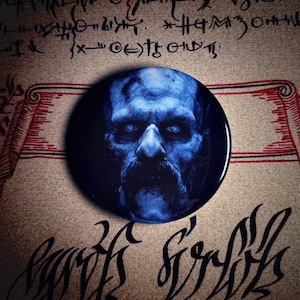 May include: A round, dark blue button featuring a detailed, blue-toned portrait of a man with a mustache and glowing blue eyes. The button is set against a backdrop of aged parchment with script and red accents.