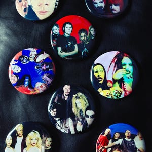 May include: A collection of round, colorful buttons featuring images of musicians and bands. The buttons display various portraits and group shots, with a variety of colors and styles. The buttons are arranged on a dark surface.