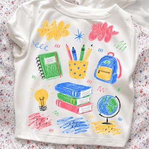 Könnte beinhalten: Weißes T-Shirt mit einer farbenfrohen Illustration zum Thema Schule. Das Design umfasst ein grünes Notizbuch, Bleistifte, Bücher, einen Rucksack, einen Globus und eine Glühbirne. Die Illustrationen sind in leuchtenden Farben wie Blau, Gelb, Grün und Rot gehalten.