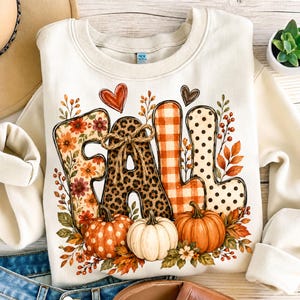Op de afbeelding: Een crèmekleurig sweatshirt met ronde hals en het woord FALL in grote, gestileerde letters. Elke letter heeft een uniek patroon, waaronder bloemen, luipaardprint, oranje ruiten en stippen. Drie pompoenen en herfstbladeren omringen de tekst.