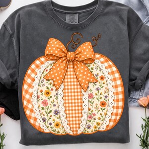 May include: A charcoal grey crewneck sweatshirt featuring a large graphic of a pumpkin. The pumpkin has alternating panels of orange gingham and a cream floral print, accented with white lace trim and a large orange polka dot bow on top.