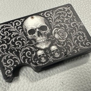 May include: Black wallet with a skull and rose design. The wallet features a large skull with a rose in its mouth, surrounded by ornate scrollwork. Two smaller skulls are also present. The design is in white against a black background.