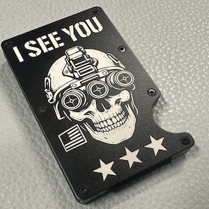May include: Black metal wallet with a skull wearing a helmet with night vision goggles. The text "I SEE YOU" is at the top. Below the skull are three white stars and a small American flag.