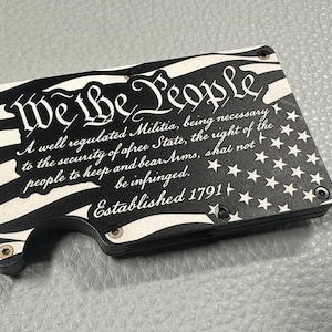 May include: Black and white wallet with the text "We The People" and a flag design. The wallet features the Second Amendment and the year 1791. The wallet is rectangular and has a curved edge.