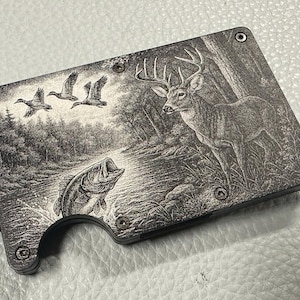 May include: A black and gray wallet with a detailed engraved scene of a buck, ducks, and a fish in a river. The wallet has a built-in bottle opener and is secured with metal rivets.