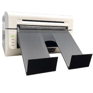 Puede incluir: Una impresora blanca y gris con bandejas de salida negras. La impresora tiene una serie de luces indicadoras en el lateral. Las bandejas de salida están inclinadas hacia abajo.