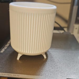 May include: A white, cylindrical planter with vertical ribbed detailing. The planter has three small, rounded feet and a wide opening. It is sitting on a gray surface, likely a 3D printer bed.