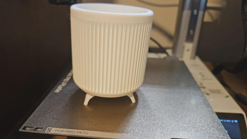 May include: A white, cylindrical planter with vertical ribbed detailing. The planter has three small, rounded feet and a wide opening. It is sitting on a gray surface, likely a 3D printer bed.
