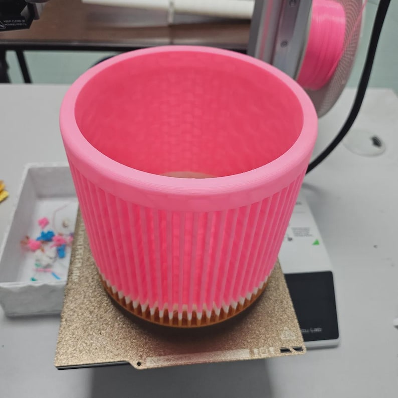May include: A bright pink, cylindrical object with vertical ribbed sides and a textured interior. The object is sitting on a gold-colored, rectangular surface. The top rim is smooth and rounded.