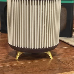 May include: A cylindrical planter with a brown rim and base, featuring vertical beige slats. The planter rests on three gold-colored legs, creating a modern, minimalist design. The planter is suitable for indoor use.