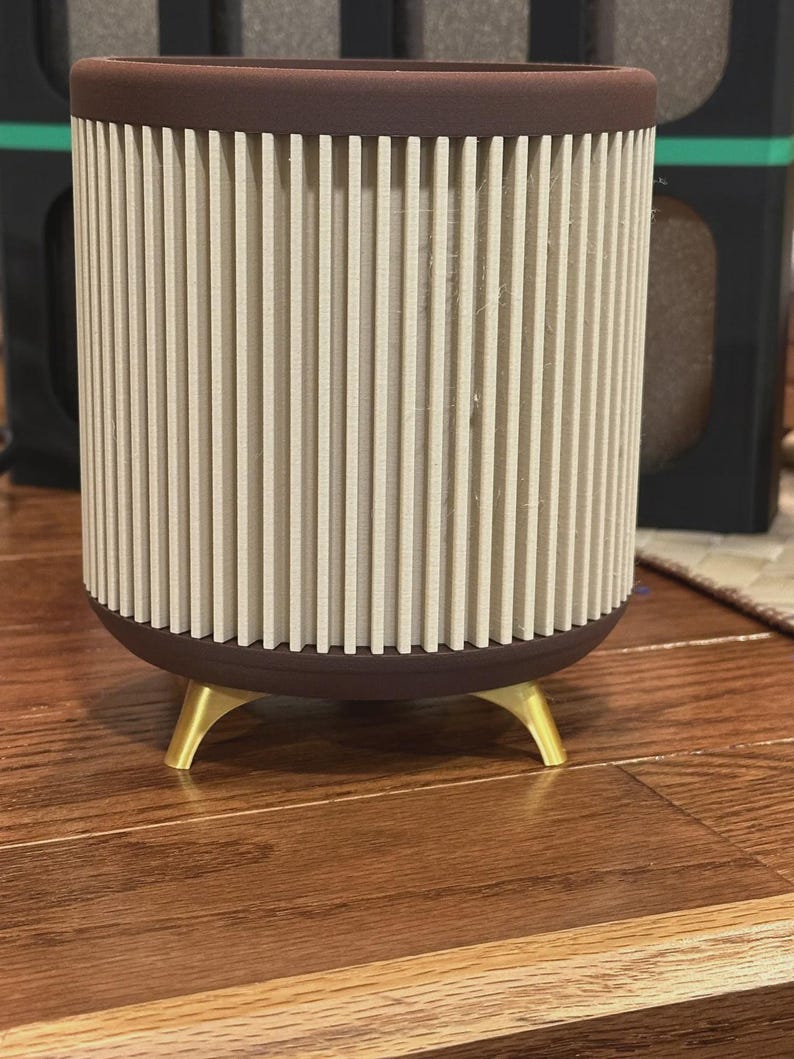 May include: A cylindrical planter with a brown rim and base, featuring vertical beige slats. The planter rests on three gold-colored legs, creating a modern, minimalist design. The planter is suitable for indoor use.