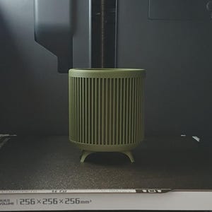 May include: A cylindrical, olive green container with vertical ribbed detailing. The container has a rounded top and sits on three small, rounded feet. The build volume is 256 x 256 x 256mm.