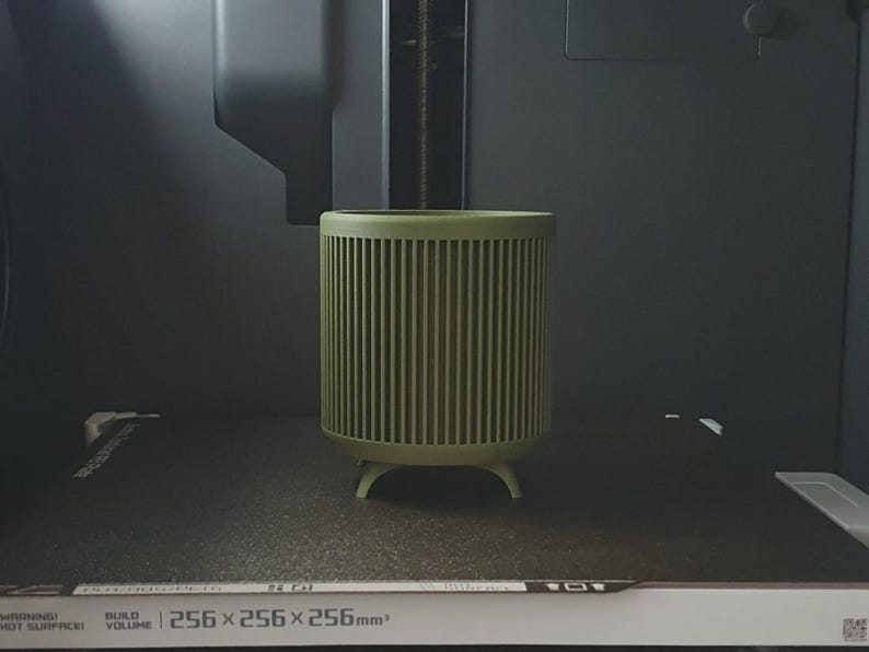 May include: A cylindrical, olive green container with vertical ribbed detailing. The container has a rounded top and sits on three small, rounded feet. The build volume is 256 x 256 x 256mm.